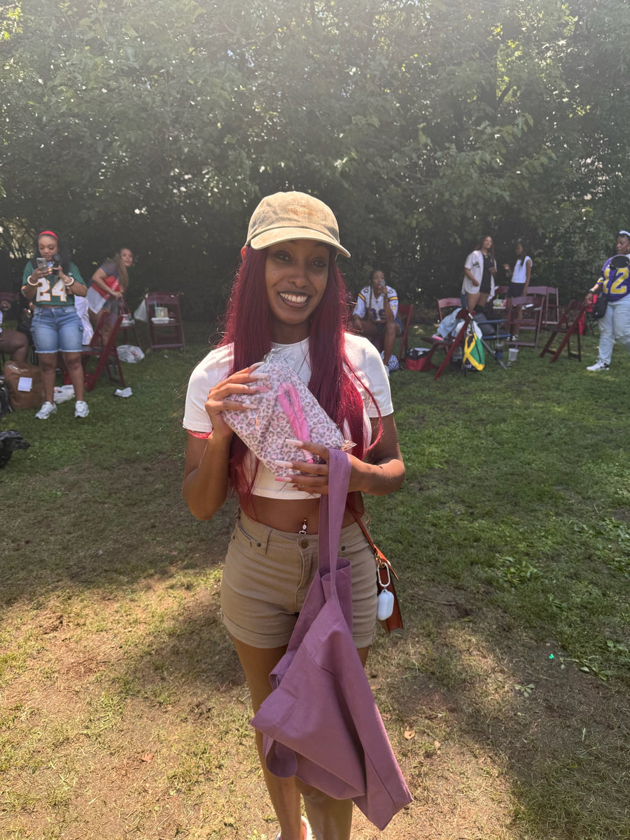 A woman with long pink hair and a beige cap stands outdoors on a grassy lawn holding a purple tote bag and a clear pouch with pink glitter. Several people and folding chairs are visible in the background among trees.